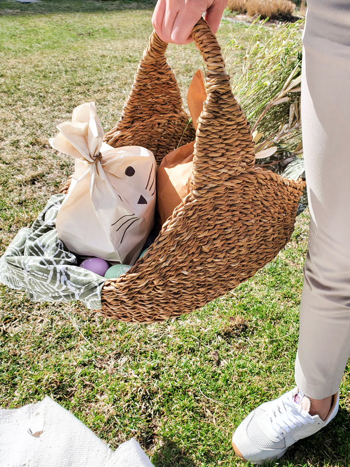 Person holding Savar picnic basket made of natural seagrass, with items inside, on a grassy field.