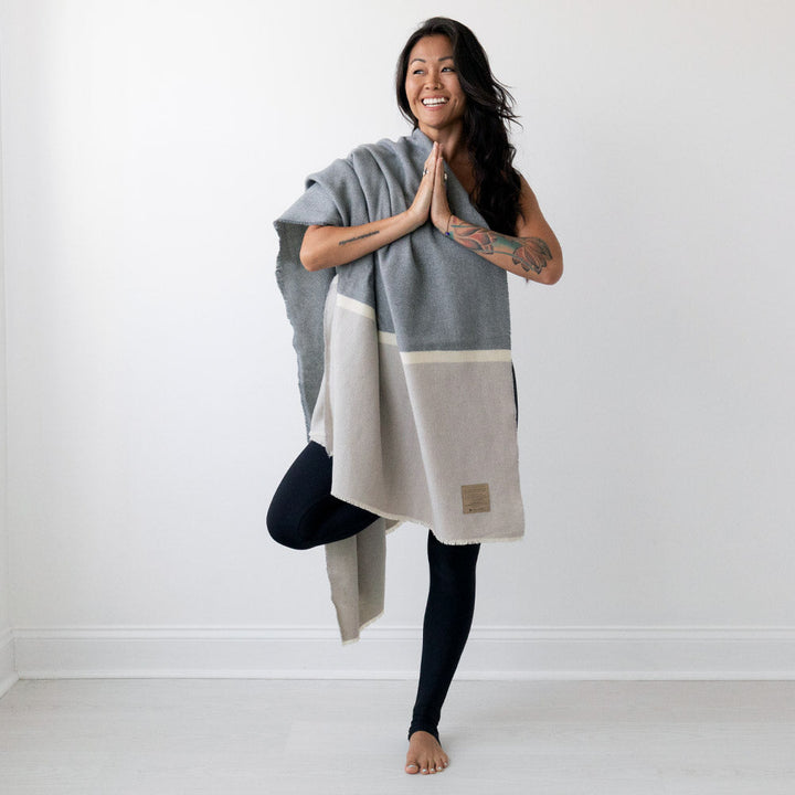 Woman wearing Namaste Blanket in neutral tones, demonstrating a yoga pose with a peaceful smile. Soft lambswool adds coziness.