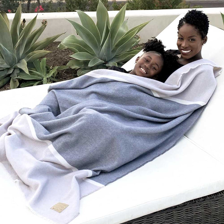 Mother and child smiling under a cozy lambswool Namaste Blanket, featuring a calming neutral palette, outdoors on a sunny day.