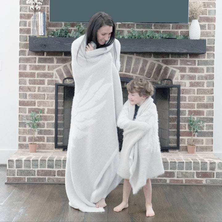 Mother and child wrapped in Dream Wings Silver Luxury Blanket by a fireplace, showcasing its soft, airy texture and winged design.