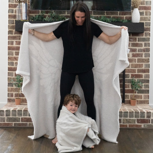 Woman and child wrapped in soft Dream Wings silver luxury blanket by a fireplace, showcasing its comfort and warmth.