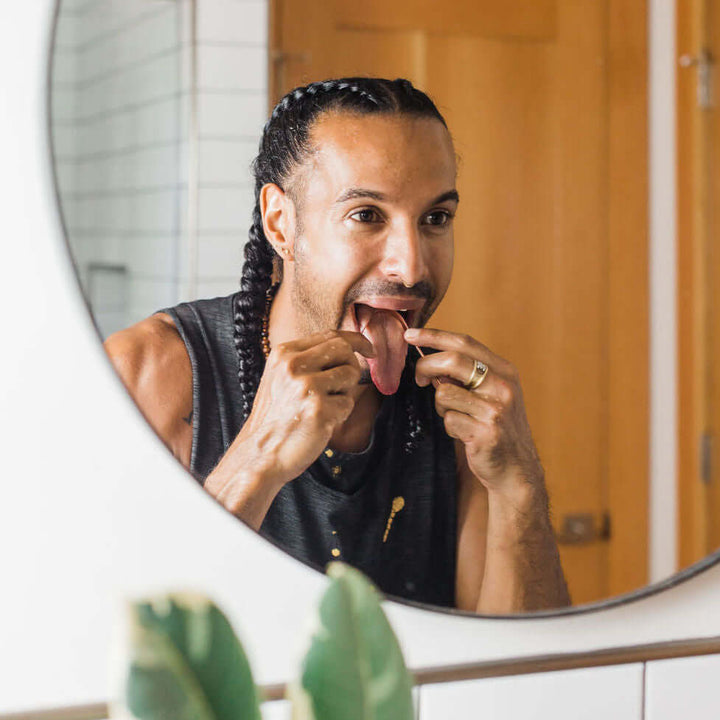 Person using a copper tongue cleaner in front of a mirror for Ayurvedic oral hygiene.