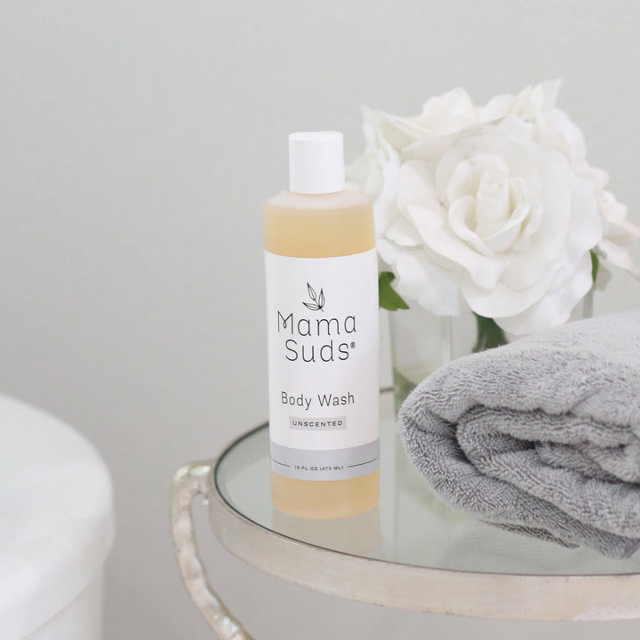 Plant Based Body Wash bottle on a glass table with white roses and gray towel, featuring natural ingredients like olive oil and essential oils.