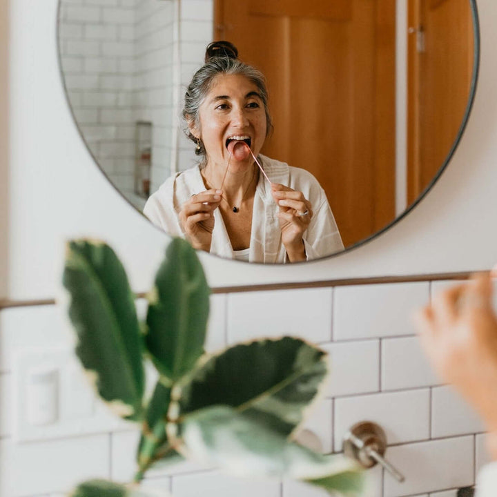 Woman using a copper tongue cleaner for daily Ayurvedic oral care in front of a mirror.