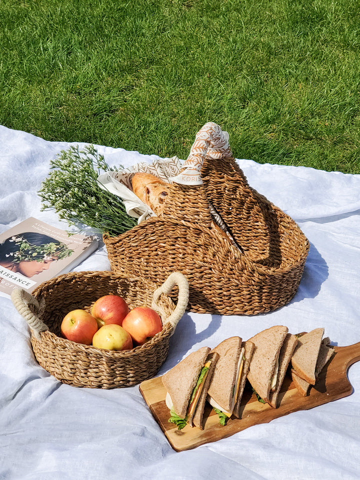 Natural Seagrass Picnic Basket with apples, sandwiches, and book on picnic blanket in grassy field