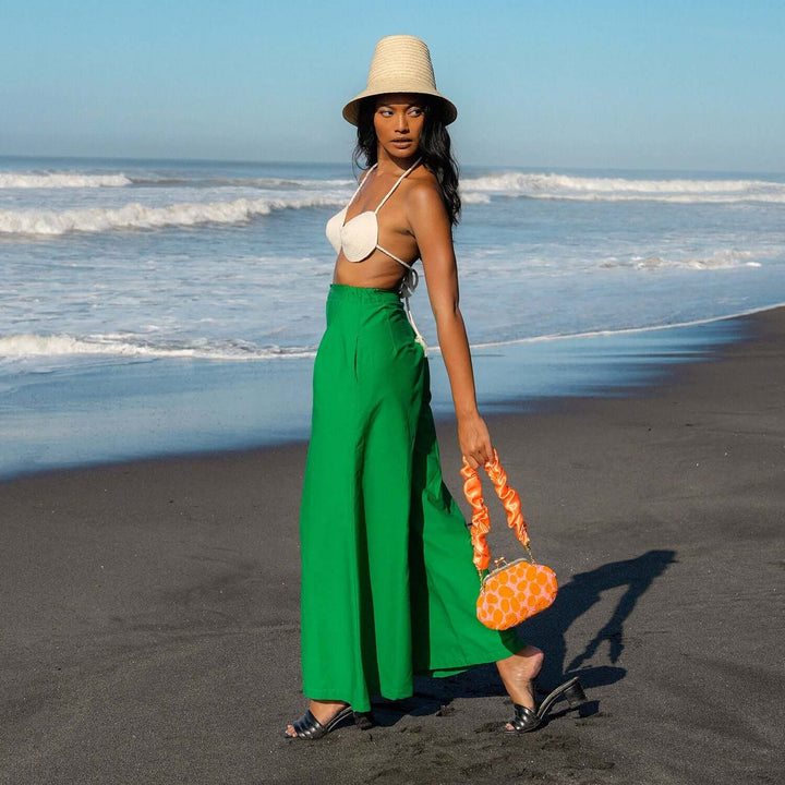 Woman wearing green Taylor wide-leg palazzo pants and straw hat, standing on a beach by the waves, holding an orange patterned bag.