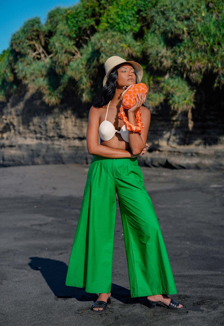 Woman wearing Taylor wide-leg palazzo pants in green, stands on a beach holding a seashell with greenery in the background.