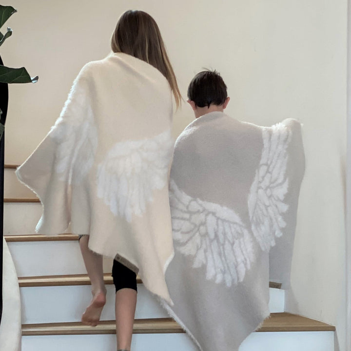 Two children wrapped in Dream Wings silver luxury blankets with feather designs, ascending stairs, showcasing soft, airy warmth.
