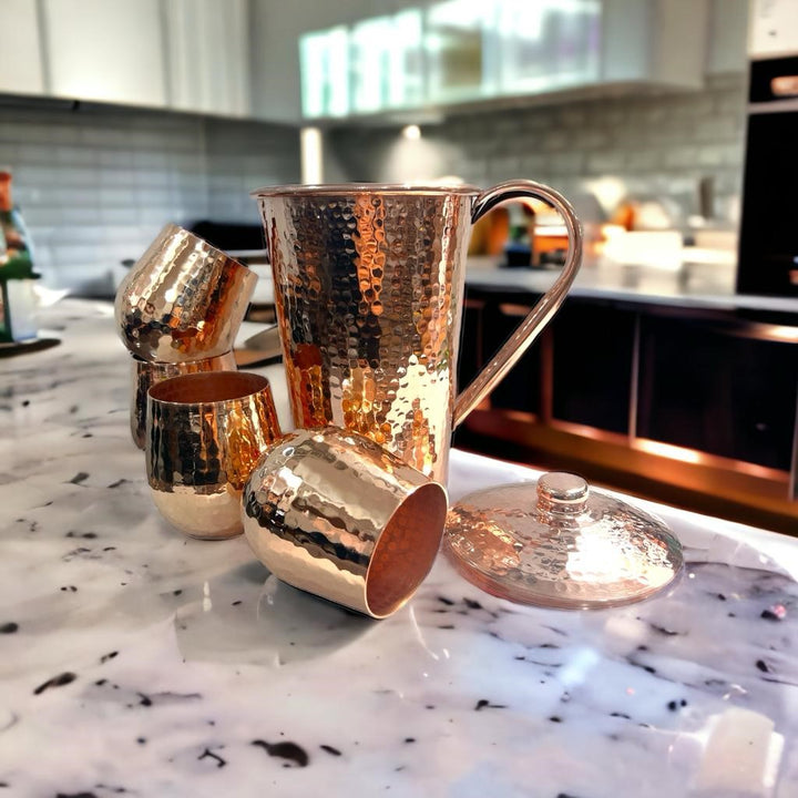 Hammered copper pitcher with lid and four tumblers on kitchen countertop, featuring a shiny finish.