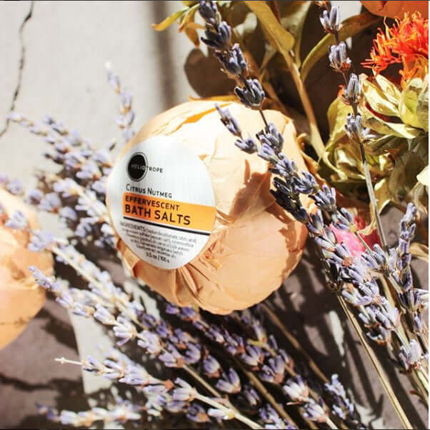 Effervescent bath salts with jojoba and silk proteins wrapped in eco-friendly paper, surrounded by lavender and flowers.