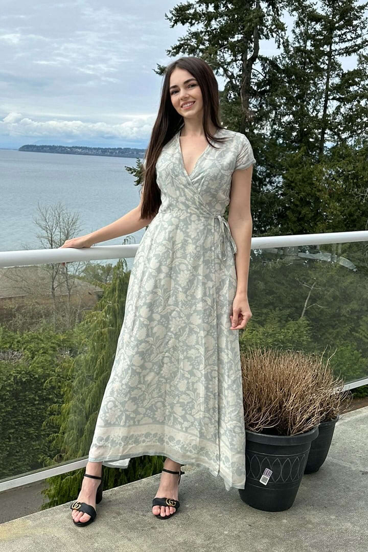 Woman wearing a blue floral maxi wrap dress, standing on a balcony with a scenic view of the ocean and trees in the background.
