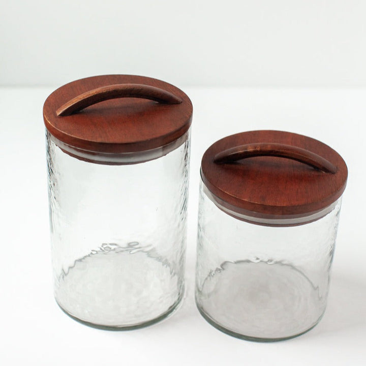 Set of clear glass canisters with hand-carved wooden lids, featuring textured design. Perfect for stylish kitchen organization. Made in India.