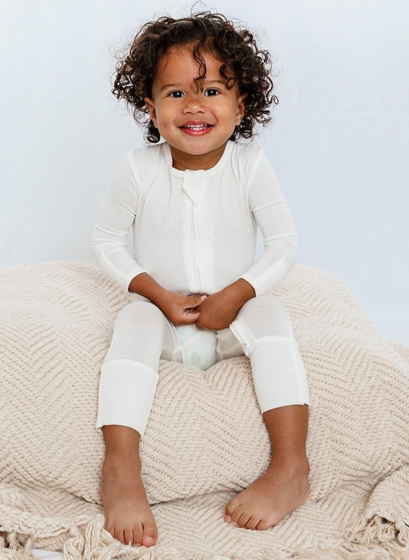 Baby wearing Luxe Bamboo Footless Romper in White with fold-over footie sitting on a soft blanket, showcasing comfort and flexibility.