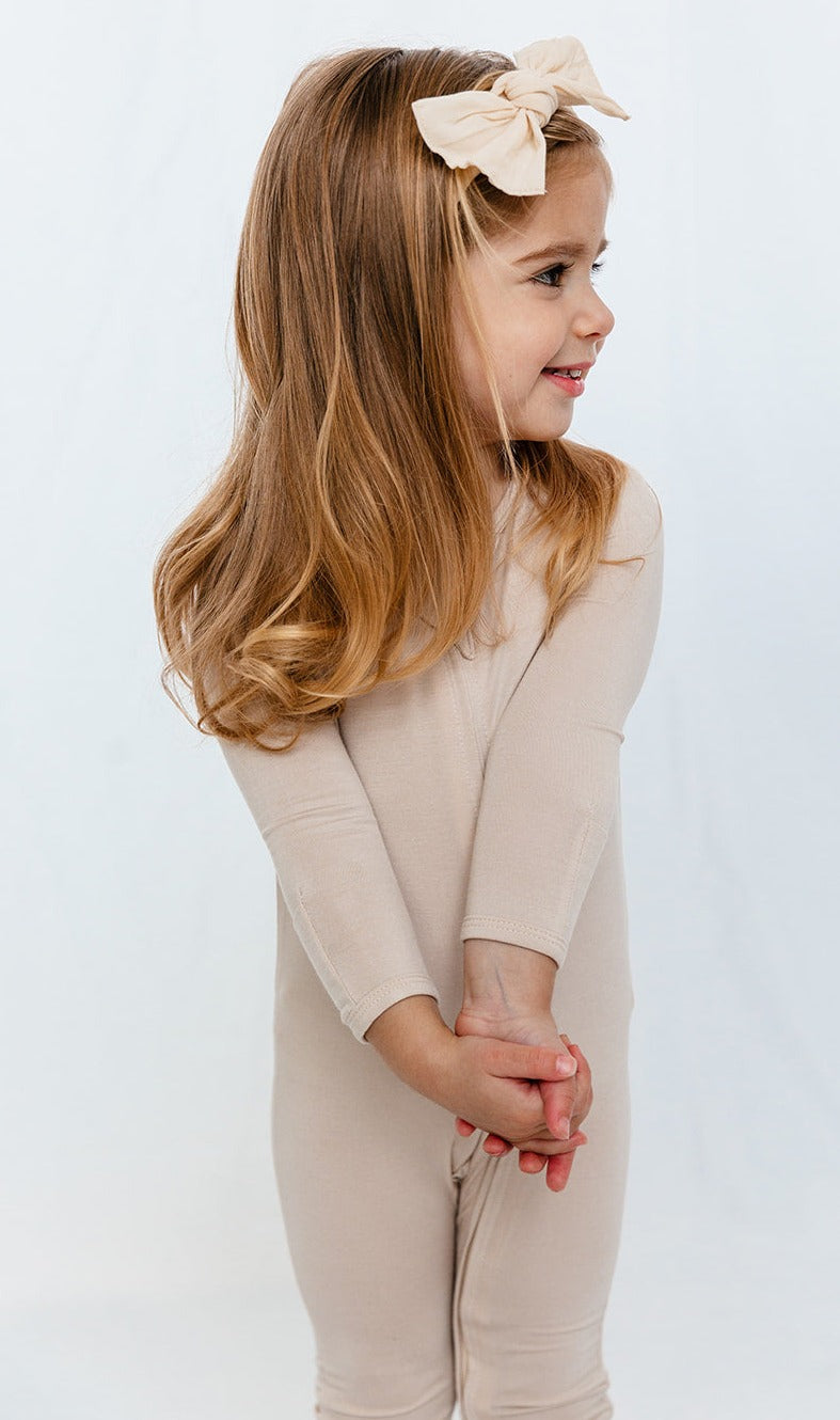 Toddler wearing Luxe Bamboo Baby Pajamas in color: Sand, showcasing soft and stretchy fabric, with a bow headband.
