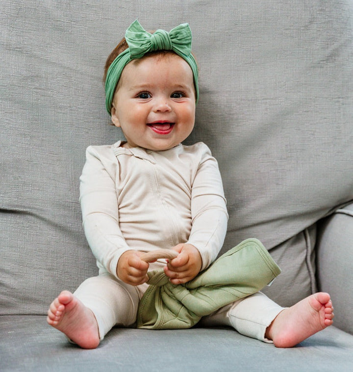 Smiling baby wearing Luxe Bamboo Footless Romper in White with green bow, sitting on gray sofa