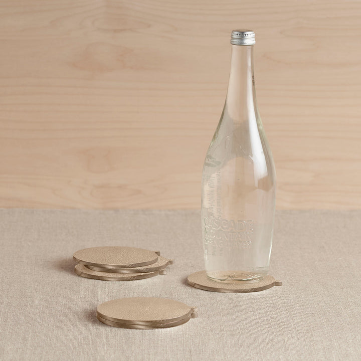 Set of six round brass coasters with decorative tabs on white marble table with glasses