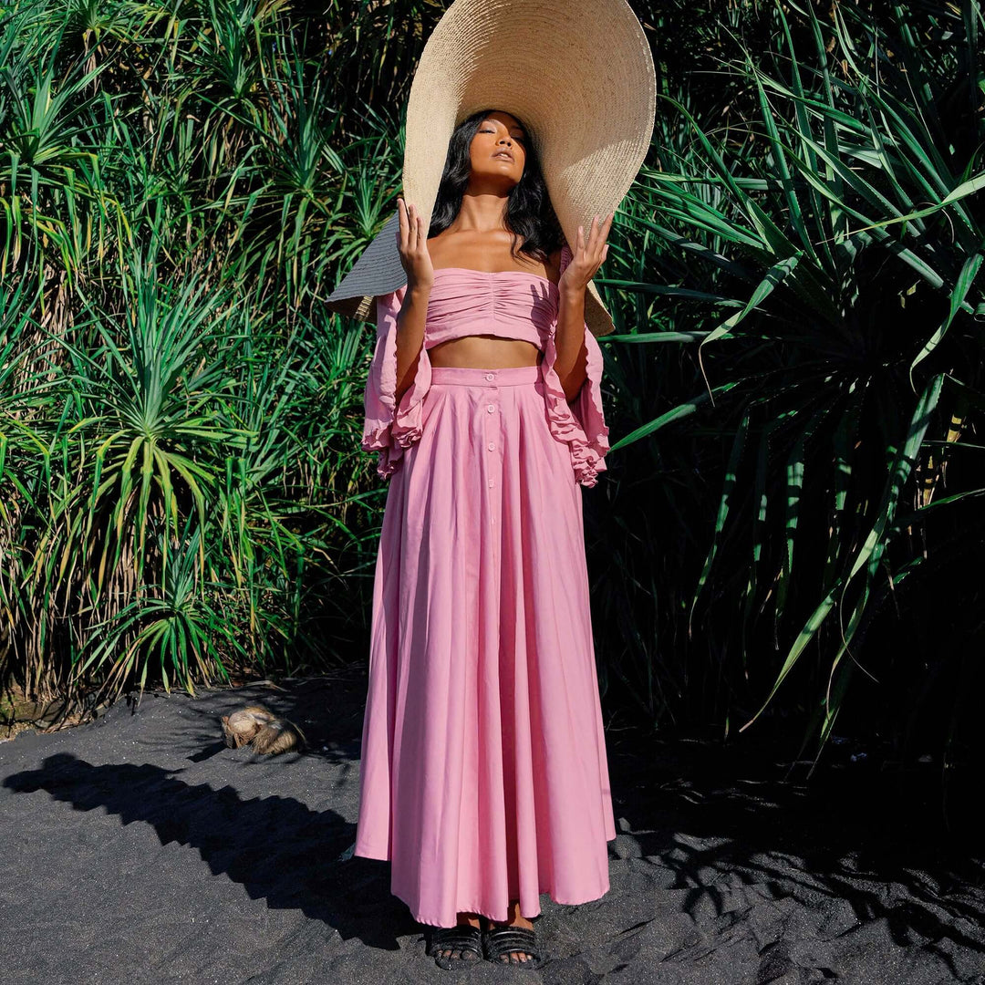 Woman wearing blush pink Arum Pleated Maxi Skirt with oversized hat, standing in nature, showcasing flowy design and elegance.