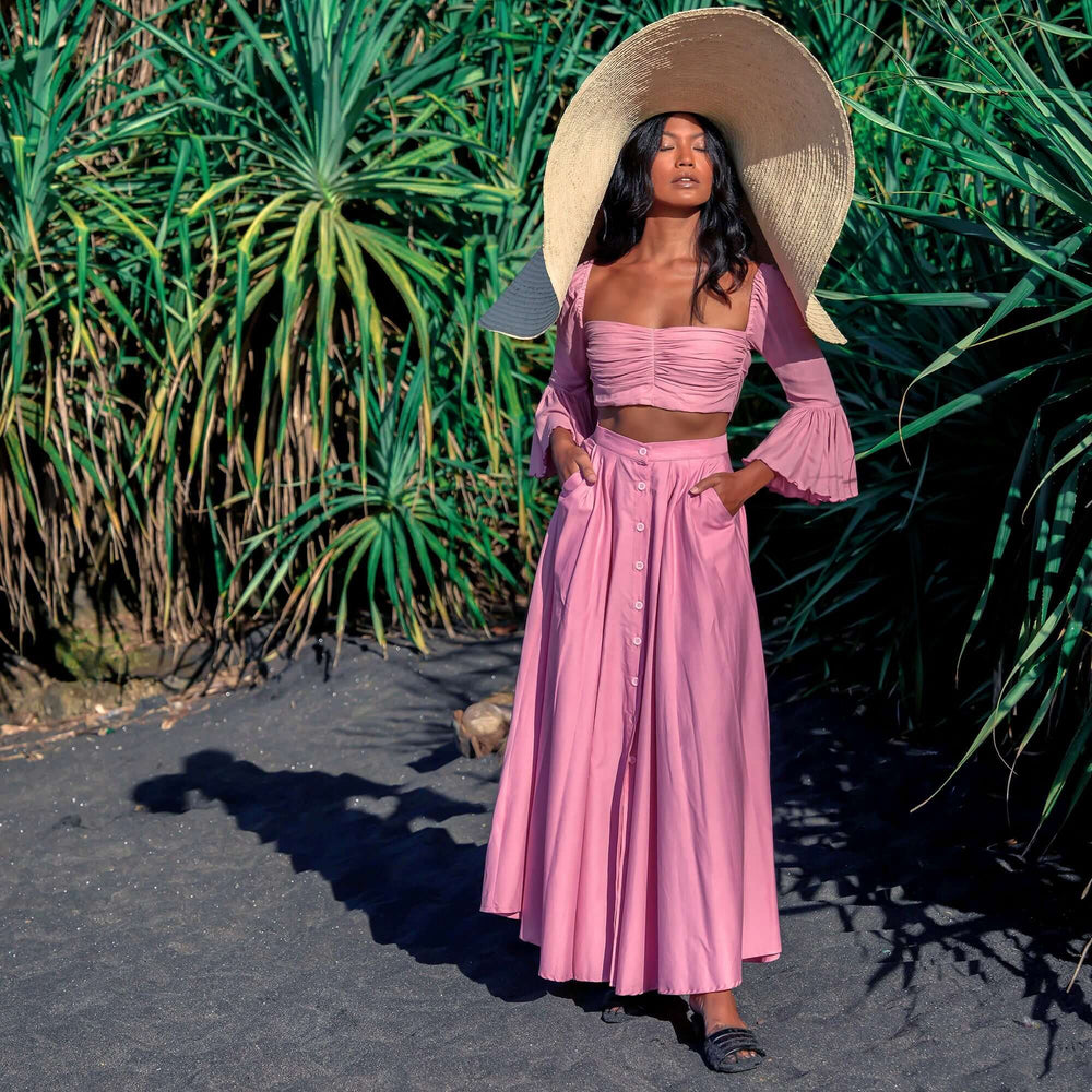 Woman wearing Arum Pleated Maxi Skirt in blush pink with a smocked waist and button-down front, standing on a beach path in a sun hat.