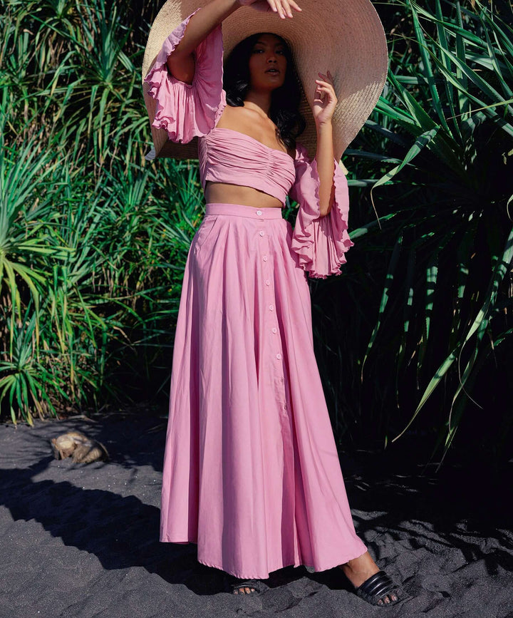 Model wearing Arum Pleated Maxi Skirt in blush pink with smocked waist and buttons, styled with wide-brim hat in lush outdoor setting.