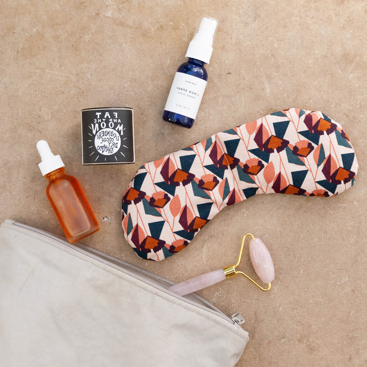 Colorful weighted eye mask with floral pattern surrounded by skincare products including a face roller, essential oils, and a small bag.