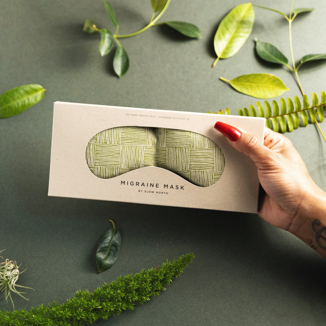 Weighted eye mask for migraine relief in packaging, surrounded by green leaves and ferns, held by a hand.