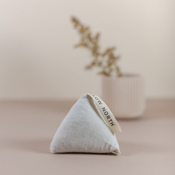 Lavender sachet in a cotton pouch for aromatherapy, displayed on a neutral background with a blurred plant in the distance.