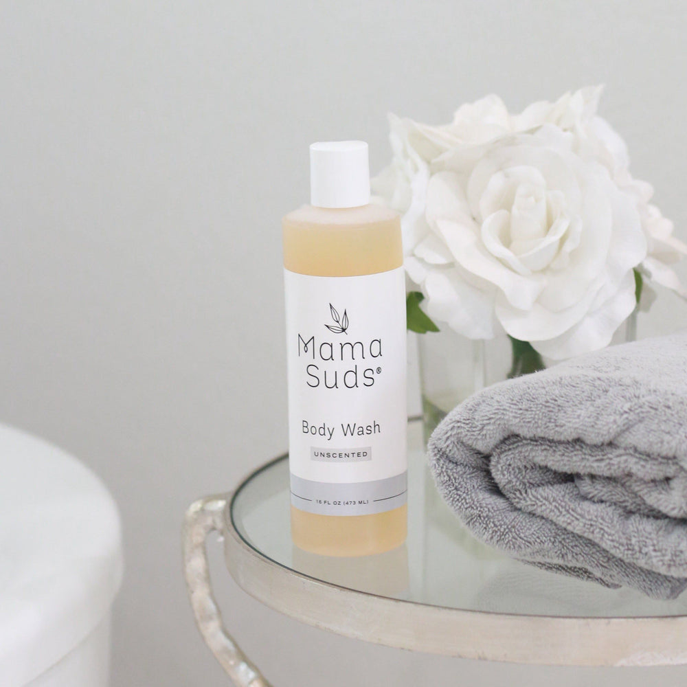 Plant Based Body Wash bottle on a glass table with white roses and gray towel, featuring natural ingredients like olive oil and essential oils.