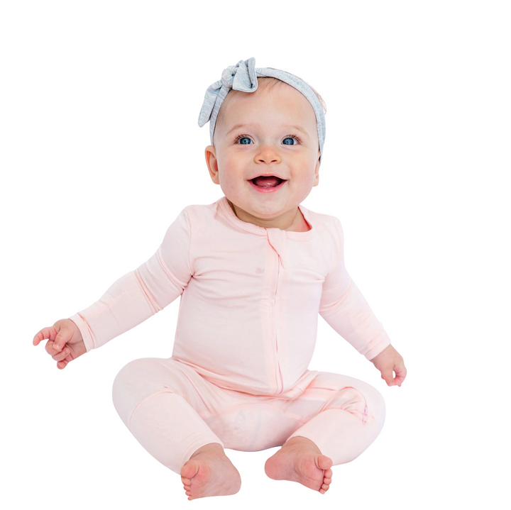 Baby wearing pink Luxe Bamboo Baby Pajamas with headband, sitting and smiling. Features soft, breathable fabric and two-way zipper for easy changes.