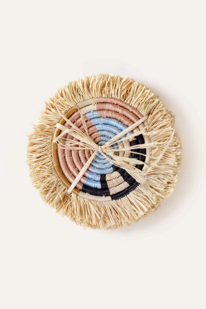 Handwoven multicolor raffia coasters with natural fringe displayed on wooden table surface