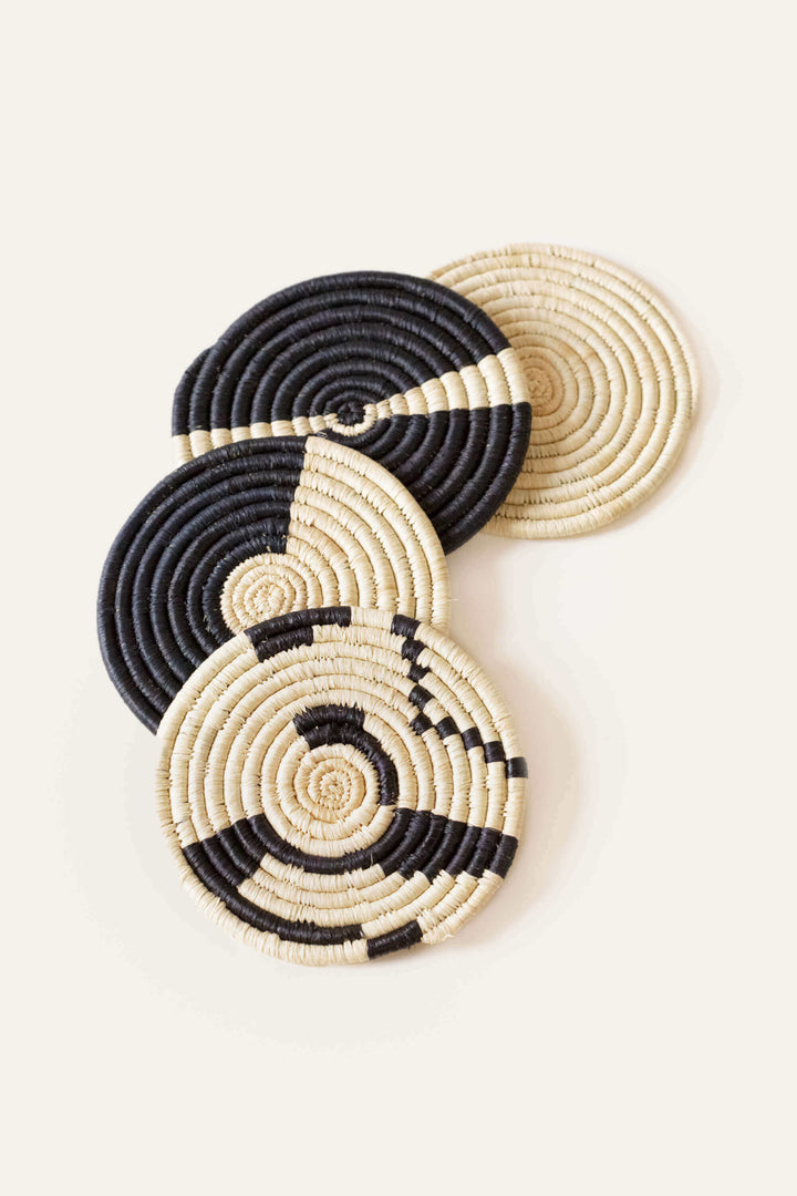 Four handwoven coasters in natural and black sisal and sweetgrass arranged on wooden table surface