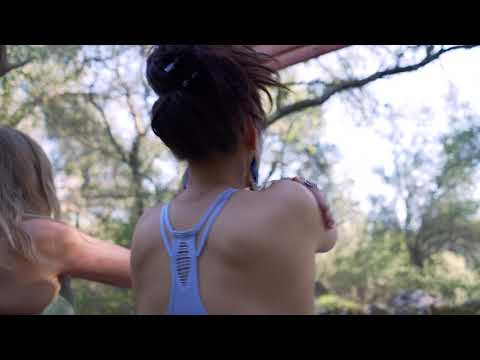 Two women practicing yoga in a forest, one wearing a blue tank top, surrounded by trees and nature.