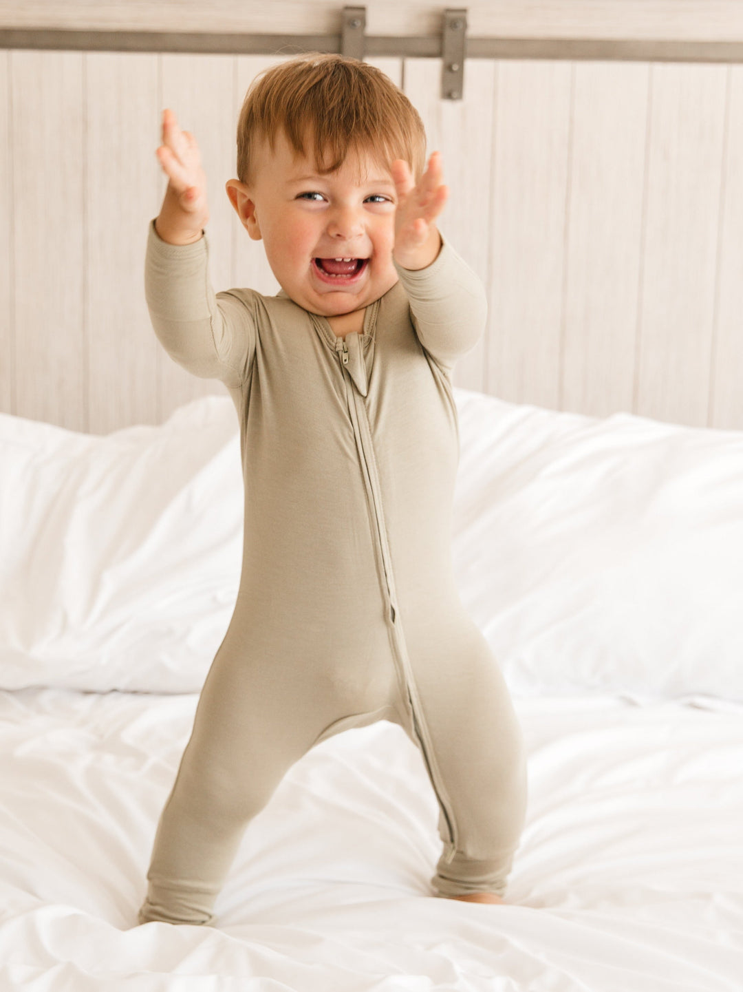 Baby wearing Luxe Bamboo Baby Pajamas in color: Sand, with two-way zipper and fold-over mittens, standing on a bed and smiling.