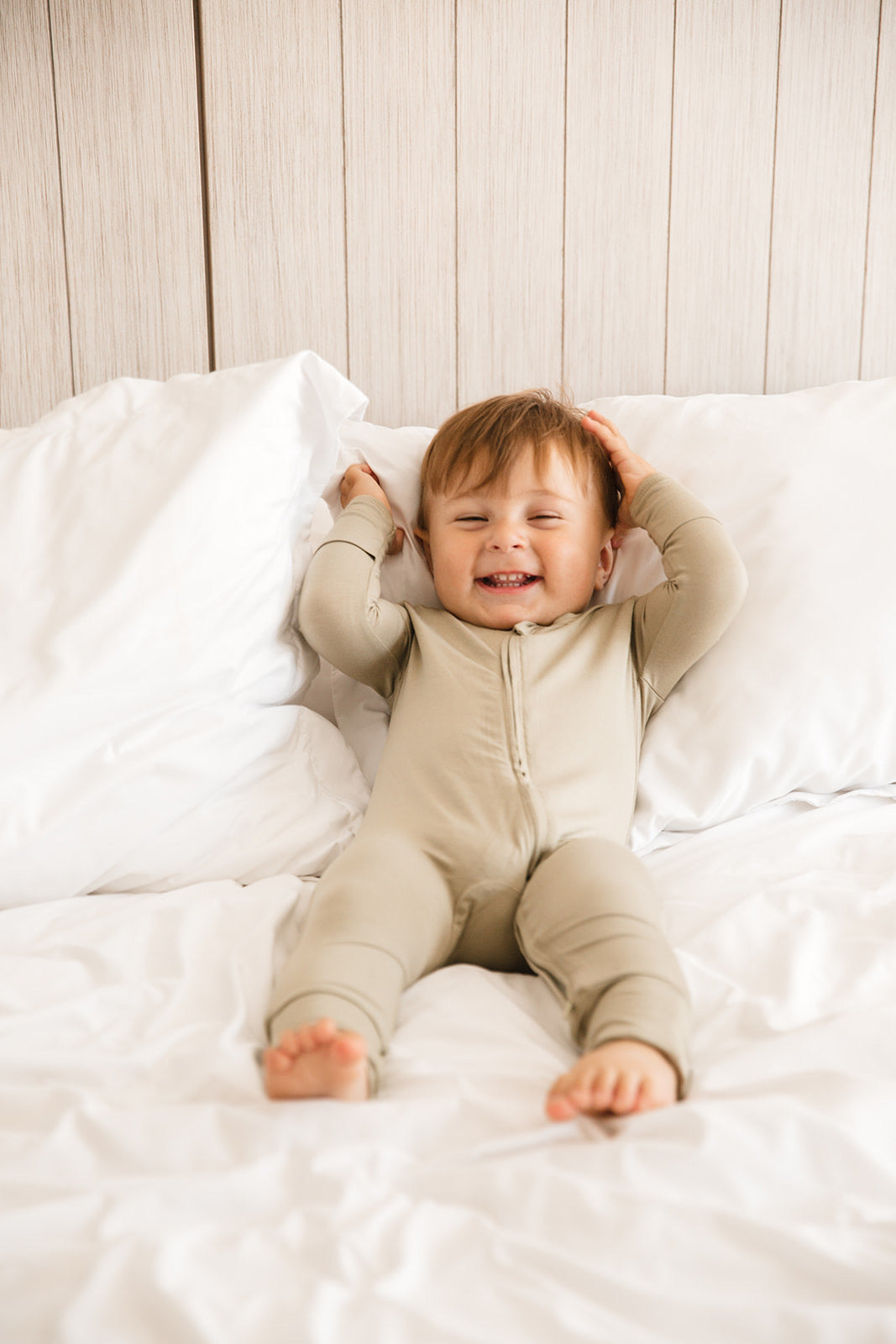 Baby wearing Luxe Bamboo Baby Pajamas in color: Sand, smiling and laying on a white bed, comfortable and soft sleepwear.