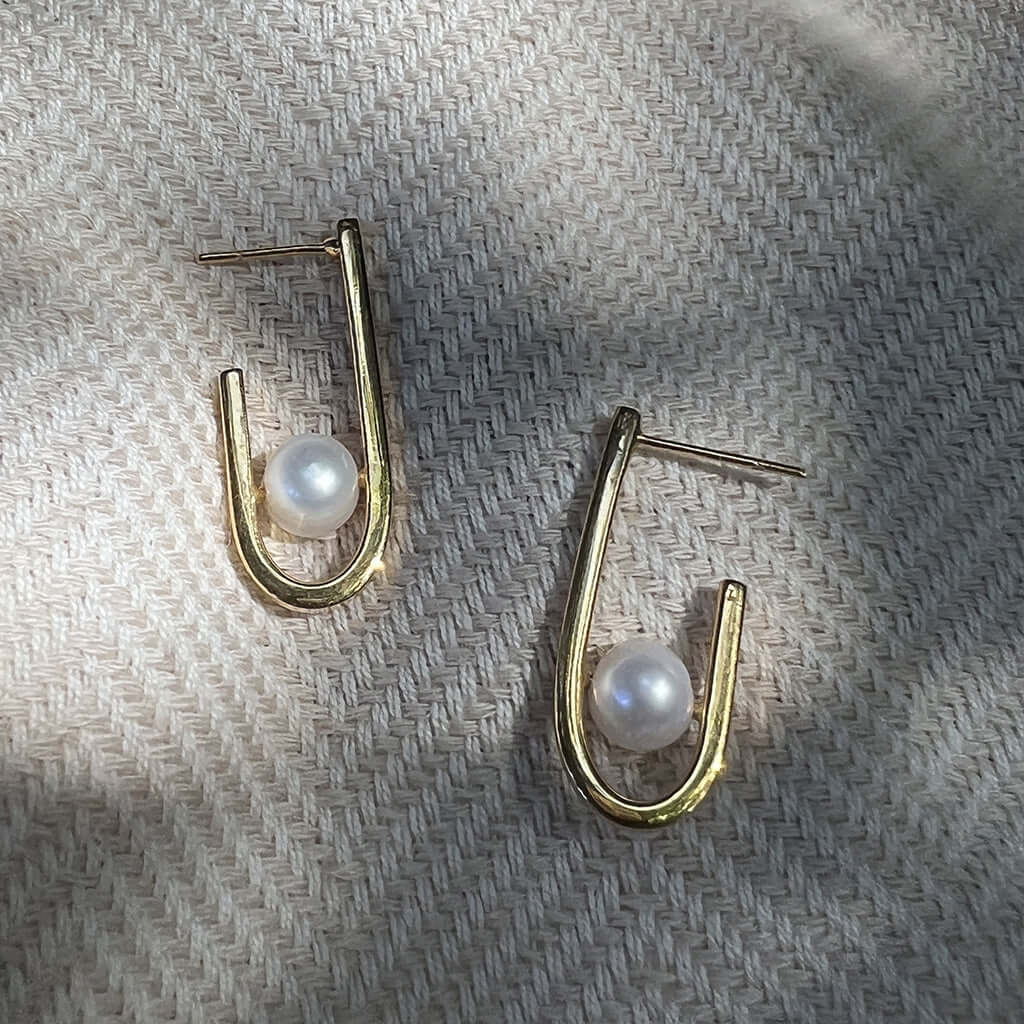 J Hoop Freshwater Pearl Earrings with gold plating, crafted from recycled bombshells, displayed on a textured surface.