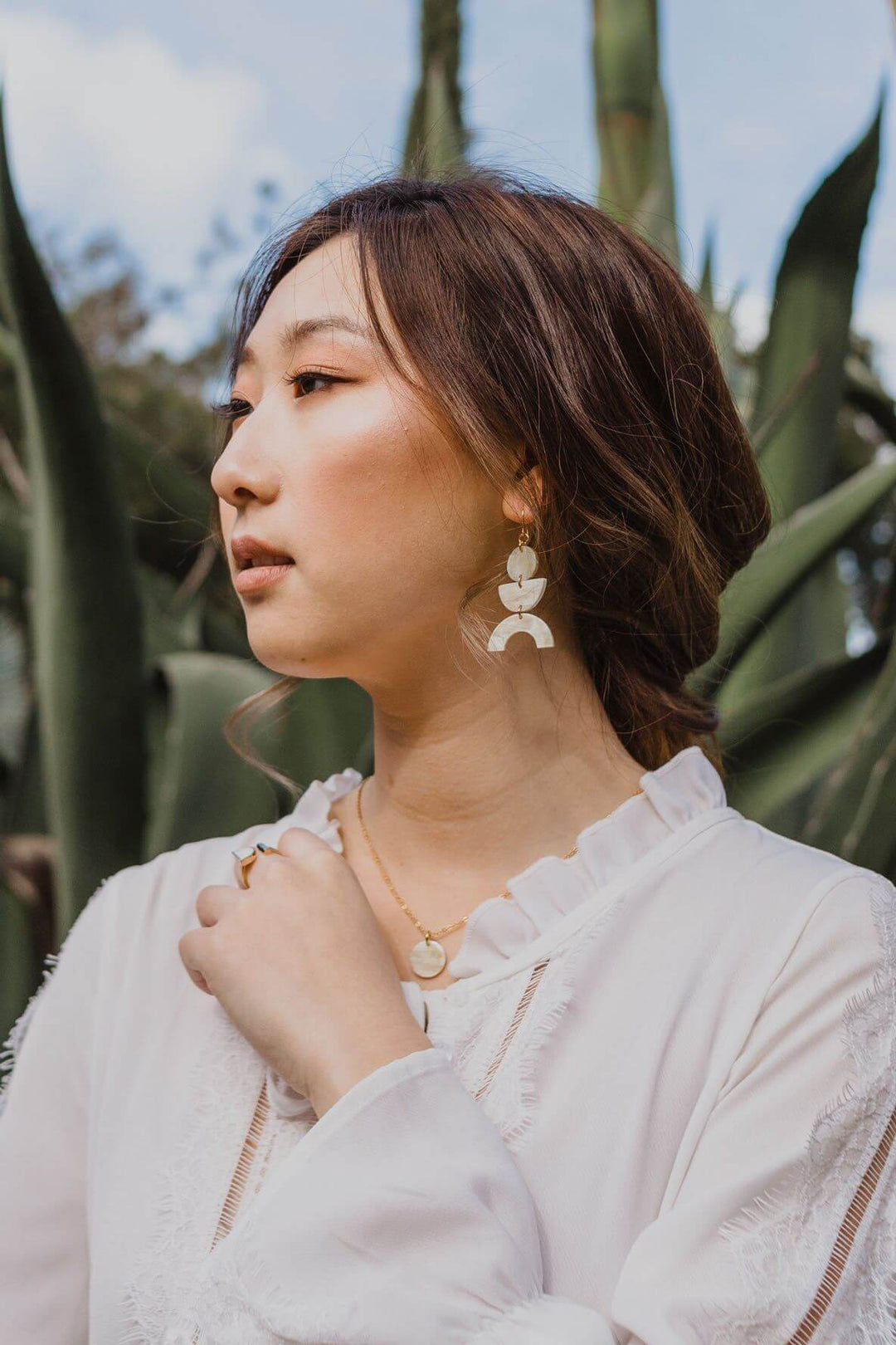 Woman wearing Vui Mung geometric buffalo horn statement earrings with gold-plated hooks, standing outdoors in a white blouse.