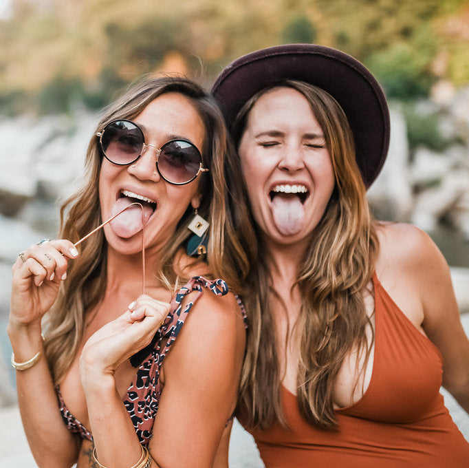 Two women practicing tongue cleaning, part of Ayurvedic oral care ritual, smiling in outdoor setting.